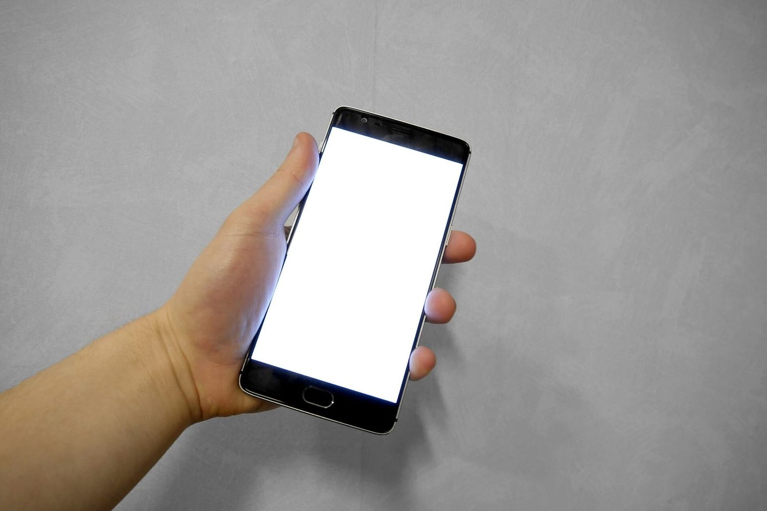 Hand hält ein Smartphone mit hell leuchtendem, leerem Bildschirm vor grauer Wand