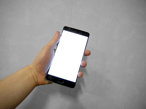 Hand hält ein Smartphone mit hell leuchtendem, leerem Bildschirm vor grauer Wand